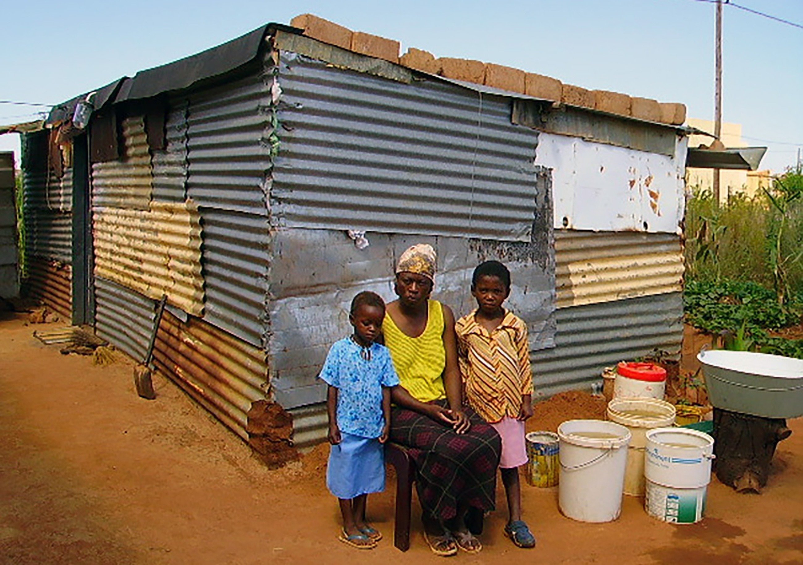 Madre africana siede con entrambi i bambini di fronte alla propria capanna in lamiera.